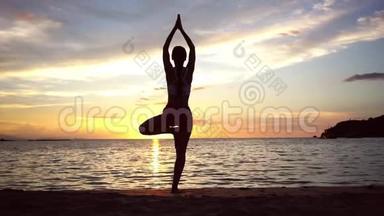 暑假里，在日落时分，女人在海滩上练习树瑜伽姿势